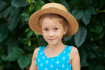 Summer holidays. A cute little girl in a summer dress and hat smiles standing among green summer foliage. Happy summer time.の写真素材