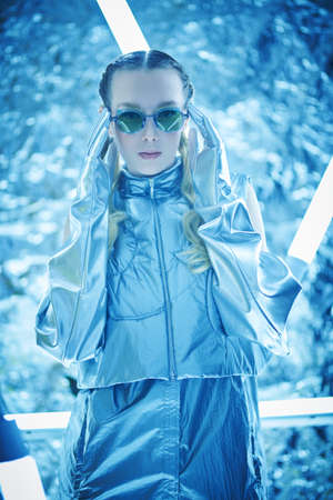 Futuristic fashion. An attractive girl with wavy blonde hair poses in trendy designer clothes and sunglasses among neon lamps. Silver background.の写真素材