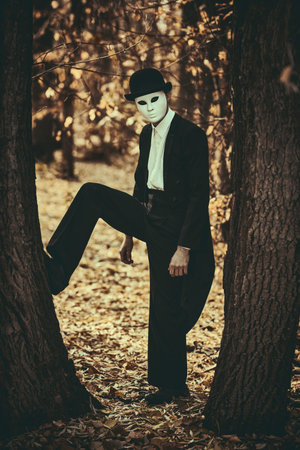 Mysterious male mime in a white carnival mask, bowler hat and black tailcoat walks in an old autumn...