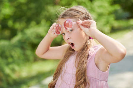 A cute girl child with blonde hair wearing a fashionable summer pink dress and sunglasses walks on a street and looks with surprise at the camera. Kid's summer fashion. Summer holidays.の写真素材