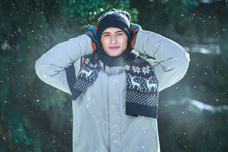 A handsome guy in a winter jacket and a knitted hat stands by snow-covered fir trees in a winter park covering his frozen ears with his mittens. Winter fashion.の写真素材