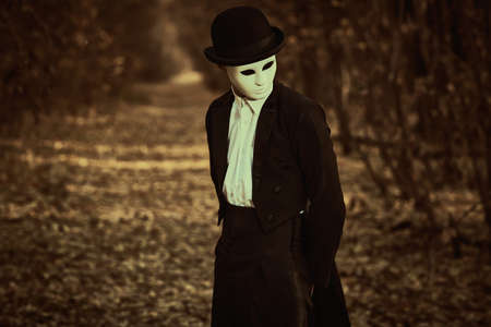 Portrait of a mysterious gentleman in a white carnival mask, bowler hat and black tailcoat standing in an old autumn park. Mime, carnival. Halloween. Vintage style.の写真素材
