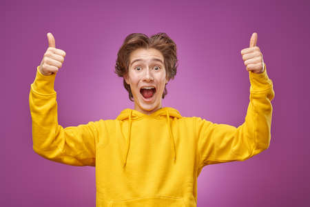 Active, happy teenager in a bright yellow hoodie rejoices emotionally and points his thumbs up. Purple background. Youth lifestyle and emotions.の写真素材