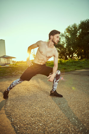Portrait of a muscular young man with bare torso doing physical exercises in the park at sunset....
