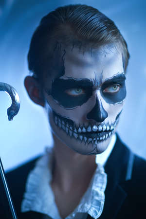 Handsome young man with a skull makeup in an elegant evening black suit, vintage white shirt and with walking stick is looking viciously away. Dia de los muertos. Day of The Dead. Halloween.の写真素材