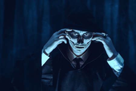 Dangerous man with skull makeup wearing a black robe with a hood stands in the moonlight against a gloomy background. Halloween. The devil, death.の写真素材