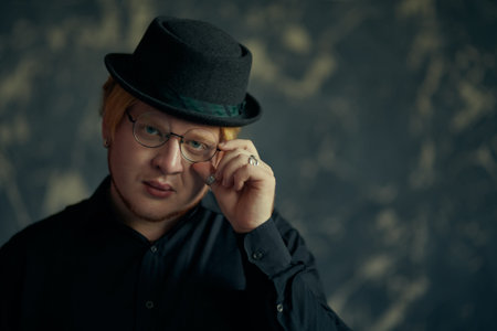A red-haired young man with chubby cheeks wearing a black shirt, stylish black hat and round glasses poses on a grunge background. Studio shot. Stylish young man. Men's style. Copy space.の写真素材
