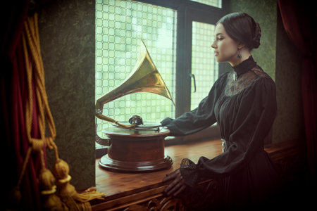 Adorable aristocratic girl in a black elegant dress with dark gathered hair stands by the window in a vintage room listening to the gramophone. The heroine of the novel, 19th century style.の写真素材