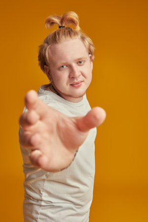 Studio shot of a plump blond guy with a high ponytail in a white t-shirt holding out his hand to the camera. Studio shot on a bright yellow background. Young people.の写真素材