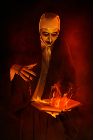An evil cursed nun with a burning bible and a cross in her hands looks at the camera on a black background. Halloween. Horrors.の写真素材