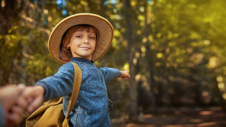 Adventure and wanderlust. An active boy in pith helmet with a backpack pulls someone's arm toward him and points with a smile forward to a wonderful summer forest. Copy space.の写真素材
