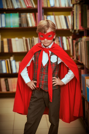 Portrait of a boy dressed in school uniform and superhero red cape and mask standing in a library among bookshelves. Super abilities in education. Children's dreams.の写真素材
