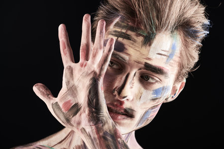 Male beauty and art. Portrait of a handsome man with colorful painted face on a black background holding hand in front of his face.の写真素材