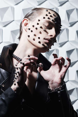 Portrait of a brutal young man in a black leather jacket and with metal spikes on his face and hand posing with closed eyes on a white tech background. Aggressive fashion.の写真素材