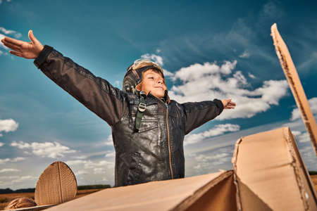 A happy little boy in a leather jacket and aviator's helmet imagines that he is flying in a cardboard plane through a blue sky. Children's games and dreams.の写真素材