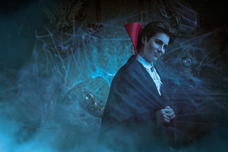Halloween. An attractive young vampire in an elegant tailcoat and a cloak stands in an old castle...