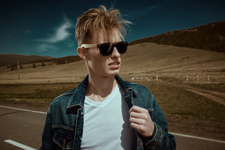 Handsome young man hitchhiker in denim clothes and black sunglasses standing on a road over picturesque landscape. Denim fashion. Road adventures, travelling.の写真素材