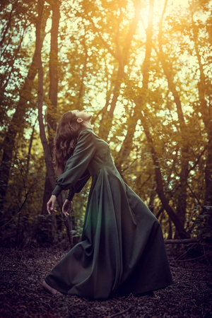 Fairy tale heroine, fantasy. A fabulous beautiful girl with wavy dark hair in a dark green long dress dances barefoot among the trees in a dense summer forest.の写真素材