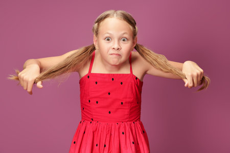 Children's emotions. A cute blonde girl in a red summer dress holds her pigtails, smiles and makes a funny face. Purple background.の写真素材