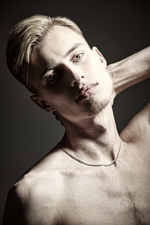 Studio shot of a handsome young man with naked torso standing calmly on a grey background with light falling on his face. Man's beauty and health.の写真素材