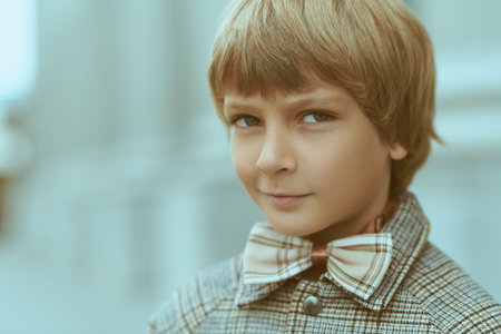 A funny fair-haired boy wearing a checkered coat and a bow tie looks with smile and distrust at the camera. Children's emotions. Street background.の写真素材