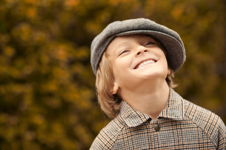 Portrait of a joyful fair-haired boy in a checkered coat and cap standing in an autumn park and...