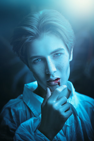 Portrait of a handsome young vampire, a teenager standing in an old castle and wiping drops of blood from his lips after biting the victim. Halloween. The hero of the vampire novel.の写真素材