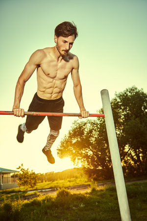 A muscular young man with bare torso is doing exercises on a horizontal bar in the park. Sports outdoors. Healthy lifestyle.の写真素材