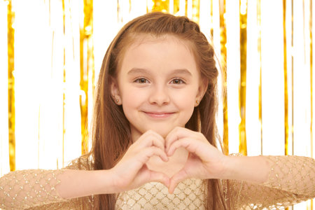 Kid's fashion and beauty. Pretty girl in a golden evening dress poses on a white background with shiny gold foil curtains showing a heart sign and smiling.  Close up shot.の写真素材