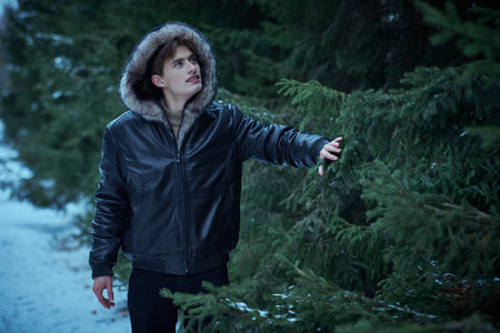 Young handsome man in stylish black leather jacket with a fur hood on his head walks in the park among fir trees on a cold winter day. Men's winter fashion.の写真素材