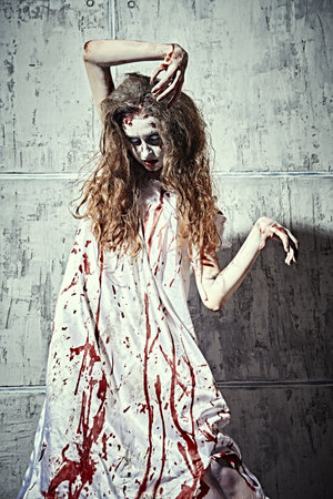 Horror scene of a creepy obsessed woman covered with blood standing by a concrete wall. Bloodthirsty zombie woman. Horror movie, thriller. Halloween.の写真素材
