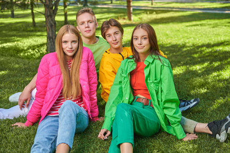 Four joyful teengers in bright youth clothes rest in a sunny park sitting on green lawn. Bright summer fashion. Youth lifestyle. Teenage age.の写真素材