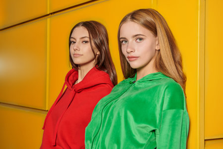 Modern youth style. Two teenage girls in bright hoodies stand leaning on a yellow wall outdoors....