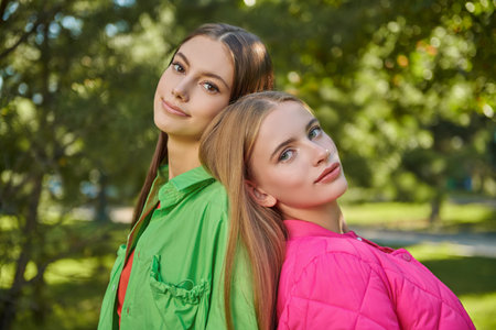 Two pretty girls teenagers in bright jackets standing in a green park. Sunny day, summer walk.の写真素材
