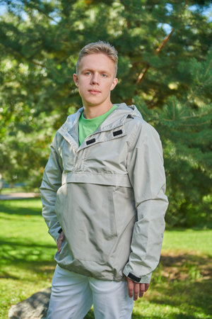 A handsome guy in stylish youth clothes stands in a green sunny park on the background of fir trees....