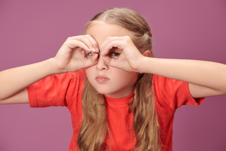 Children's emotions and games. A funny blonde girl with pigtails in a red t-shirt makes a funny face and looks with suspicion through imaginary binoculars. Purple background.の写真素材