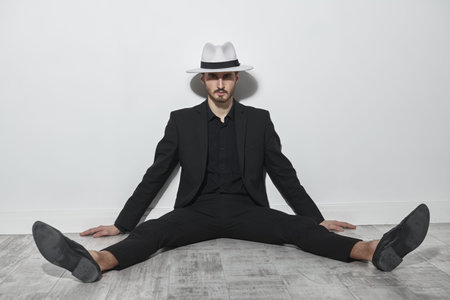Men's fashion. An elegantly dressed brunet man in a black suit and white hat sits on the floor leaning against a white wall. Full-length studio portrait.の写真素材