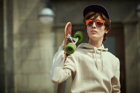 A modern teenager boy in a hoodie, hat and sunglasses is standing on the street with a longboard in his hand. Activity, summer and youth lifestyle. Copy space.の写真素材