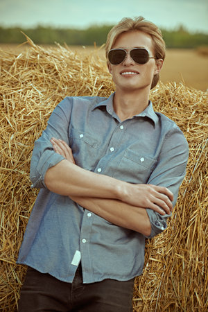 Fashion shot. Portrait of a handsome male model with blond hair wearing fashionable sunglasses and denim shirt posing by a haystack against an autumn landscape. Glasses fashion.の写真素材