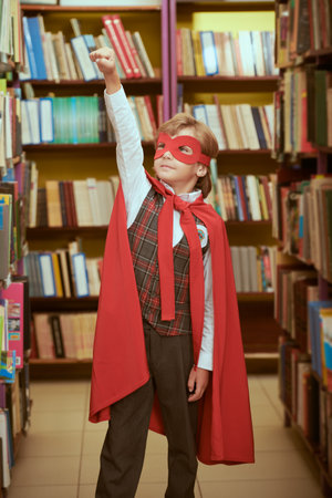 Super kid. A boy dressed in school uniform and superhero red cape and mask stands in a library by bookshelves holding one arm up. Super abilities in education. Children's dreams.の写真素材