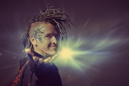 A mature male shaman in ethnic clothes stretches his hand forward with a smile, inviting you into his mysterious world. Dark background with mystical light. Halloween, Fantasy. Copy space.の写真素材