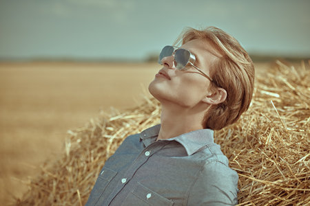Fashion shot. Portrait of a handsome male model with blond hair wearing fashionable sunglasses and denim shirt relaxing by a haystack against an autumn landscape. Glasses fashion.の写真素材