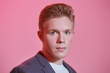 Close portrait of an elegant young man in a suit looking carefully at the camera. Pink background. People concept.の写真素材