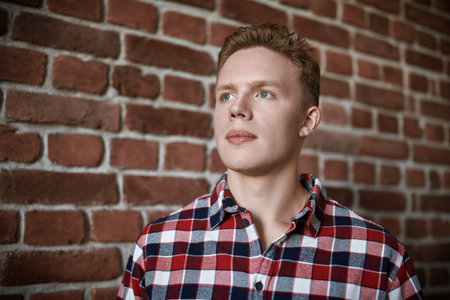 Portrait of a handsome modern guy dressed in a chekered shirt in a loft style room on the background of a brick wall. Young people, lifestyle.の写真素材