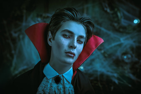 Traditional vampire. Portrait of a teenage vampire aristocrat in an elegant suit and cloak with a red collar, who looks at the camera with drops of blood on his lips. Halloween.の写真素材