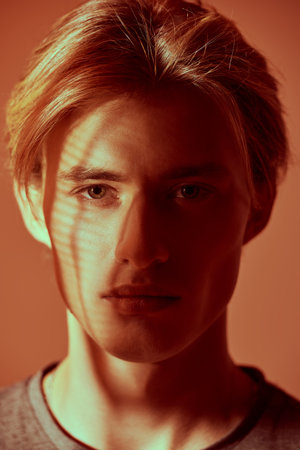 Male beauty. Art shot of a handsome blond man with an ornament of shadows on the face on an orange...