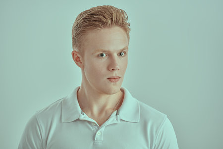 Portrait of a young blond man in a white polo shirt standing with a thoughtful expression on his face. White background. People, emotions.の写真素材