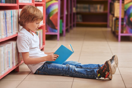 Education. A cute boy in casual clothes sits on the floor in the library reading a book. Modern children.の写真素材
