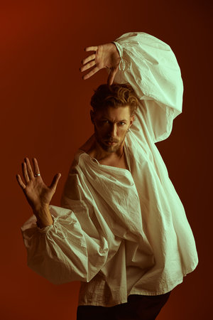 A handsome red-haired mature man in a white vintage shirt with wide sleeves poses on a burgundy...