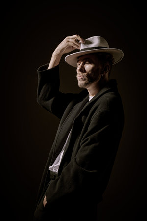 Dark key portrait of a well-groomed mature man with beard and moustache posing in a black coat and a white hat on a black background. Male beauty and fashion. Noir style.の写真素材